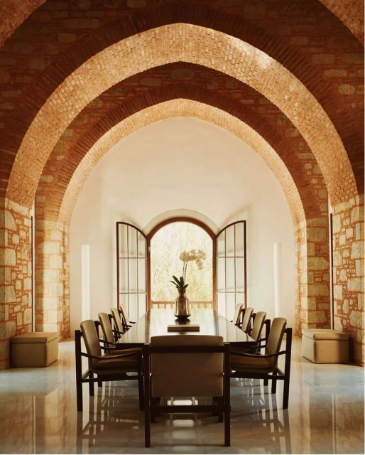 Indoor dining area featuring a long table with chairs, brick arches, and an open window, creating a bright and inviting space.