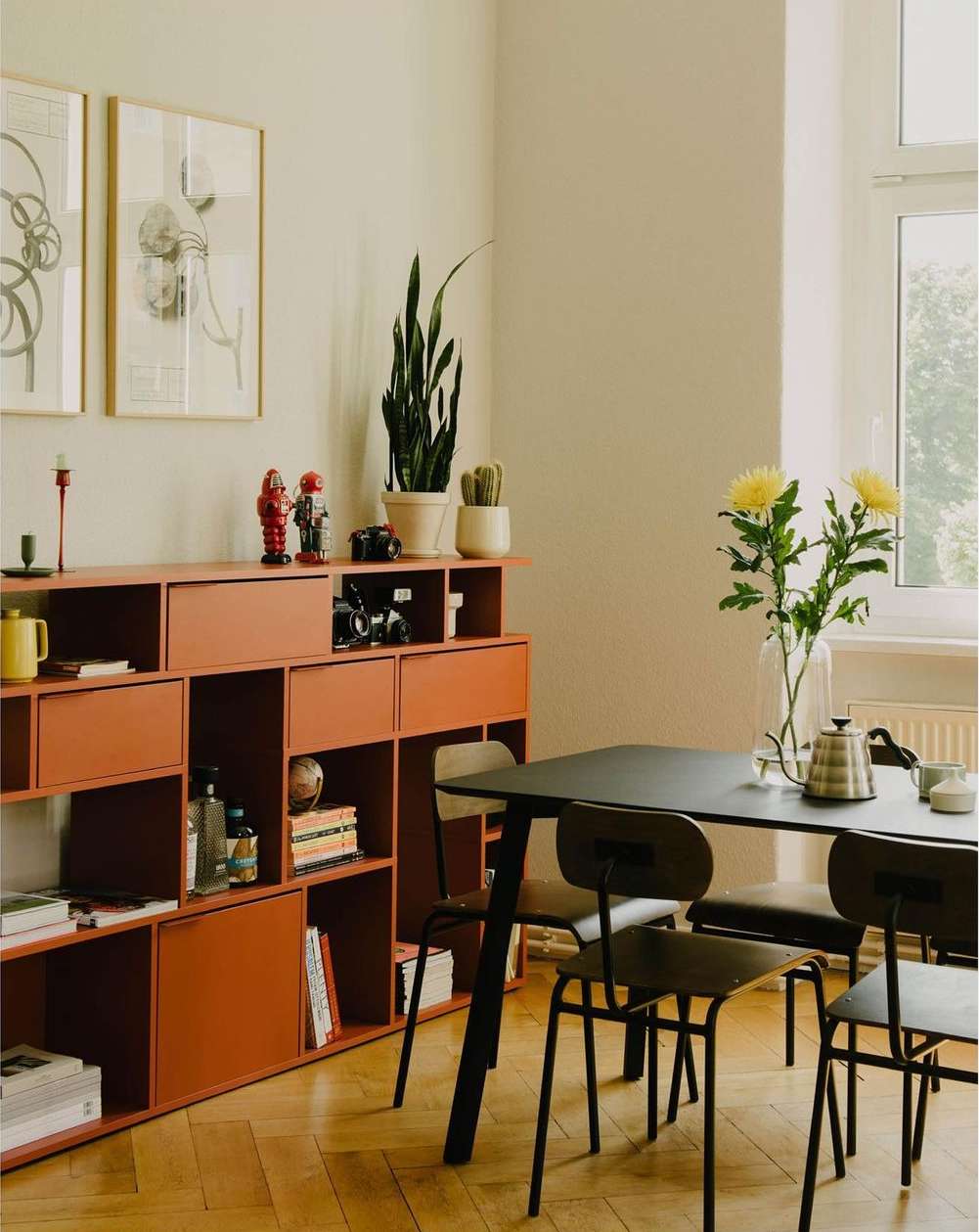 Interior of a dining room with a black table and chairs, a coral colored shelving unit, artwork, plants, and a herringbone floor.
