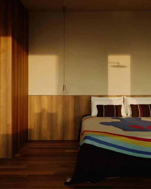 Sunlit bedroom interior featuring a bed with white pillows, striped blanket, and wooden wall paneling, illuminated by natural light.