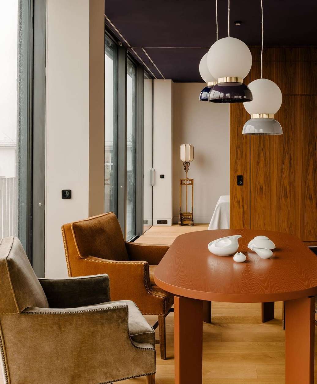 Interior view showcases a modern dining room with brown chairs around an orange table, pendant lights, and large windows.