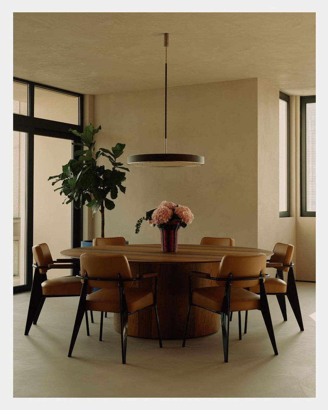 Interior of a dining room with a round wooden table surrounded by six leather and black chairs, underneath a modern pendant light.