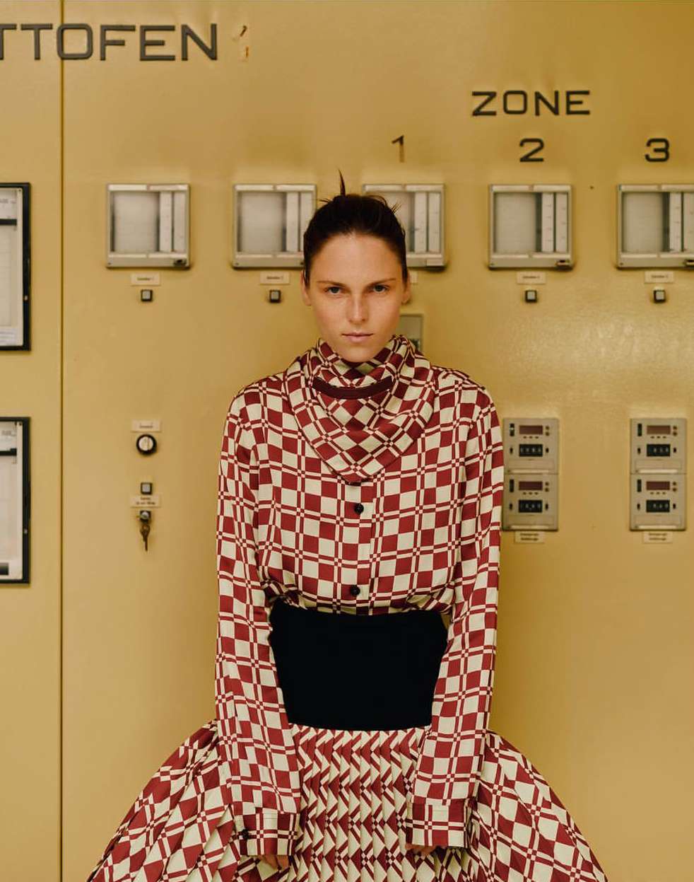 Fashionable adult woman in a checkered long-sleeved top and skirt poses indoors in front of a large industrial control panel.
