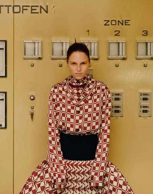 Fashionable adult woman in a checkered long-sleeved top and skirt poses indoors in front of a large industrial control panel.