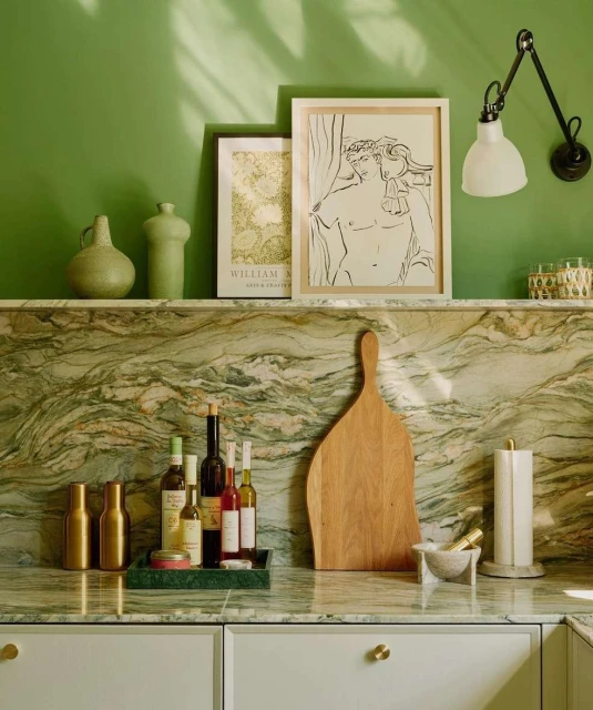 Interior setting features kitchen countertop with marble backsplash, assorted bottles, art prints, and decorative objects against green wall.
