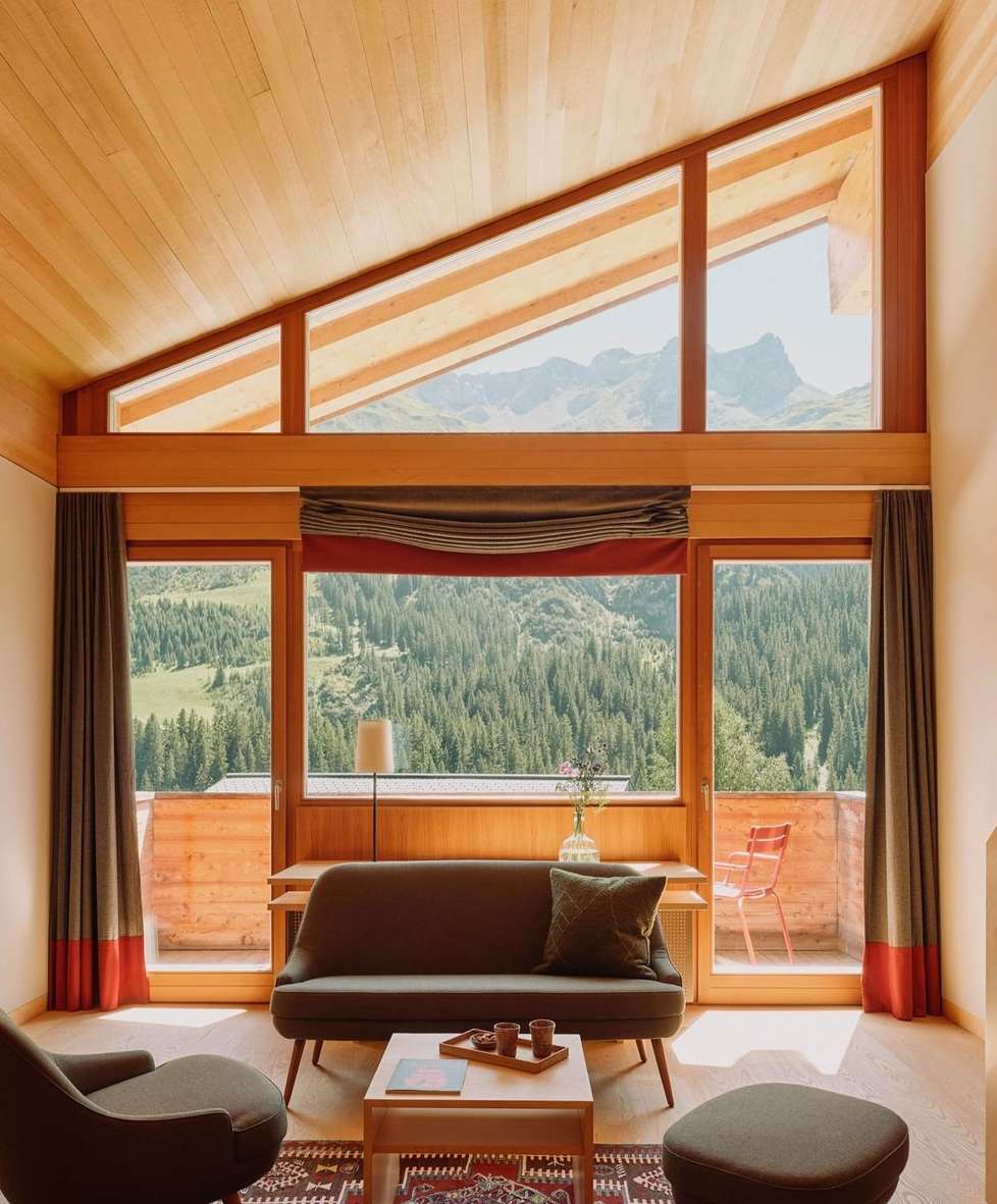 Warm interior features a grey sofa and armchair, a wood ceiling with triangular window, and a mountain view in the background.