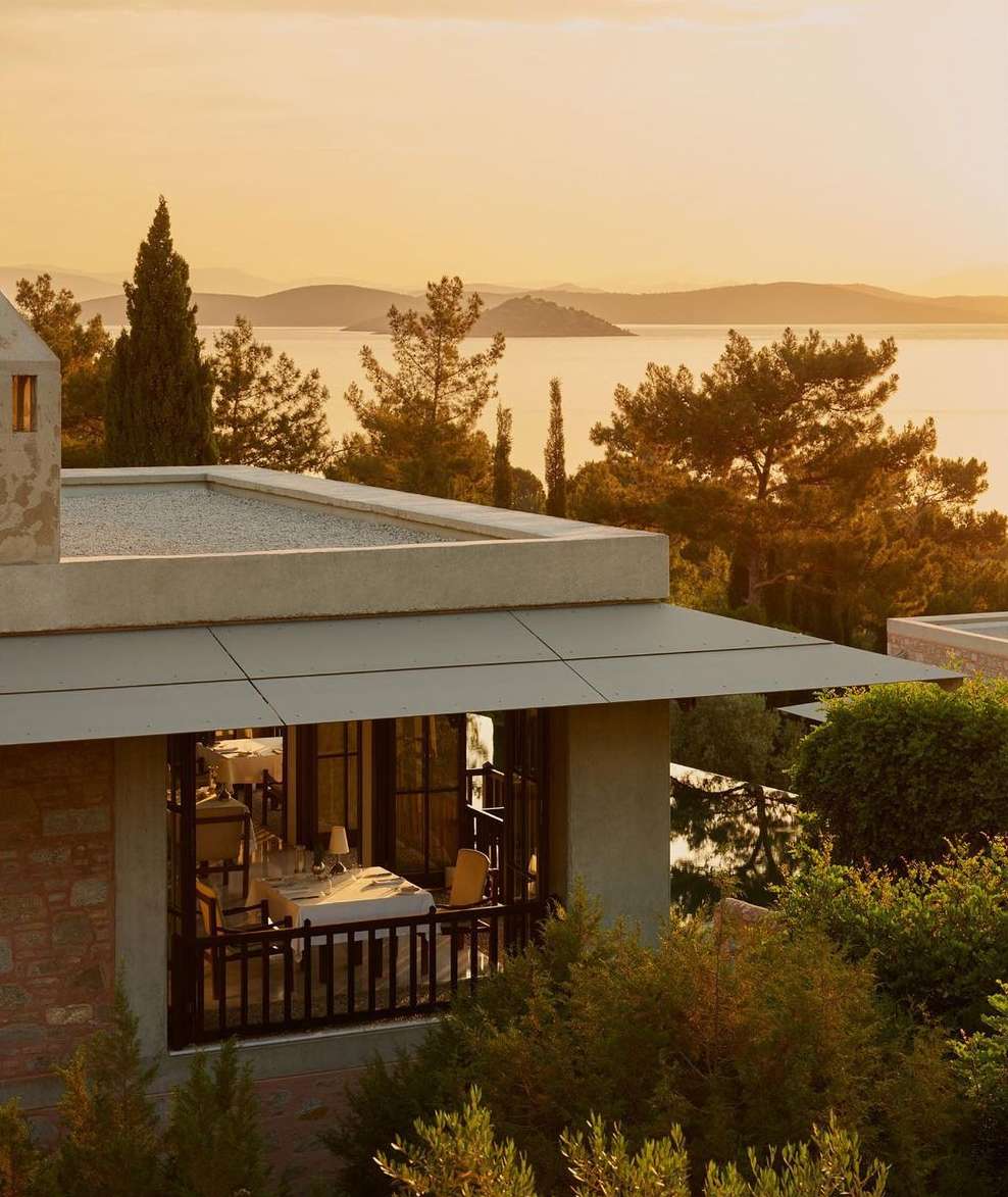 Exterior view of a restaurant with outdoor seating overlooking the ocean, featuring stone architecture and surrounding trees during sunset.