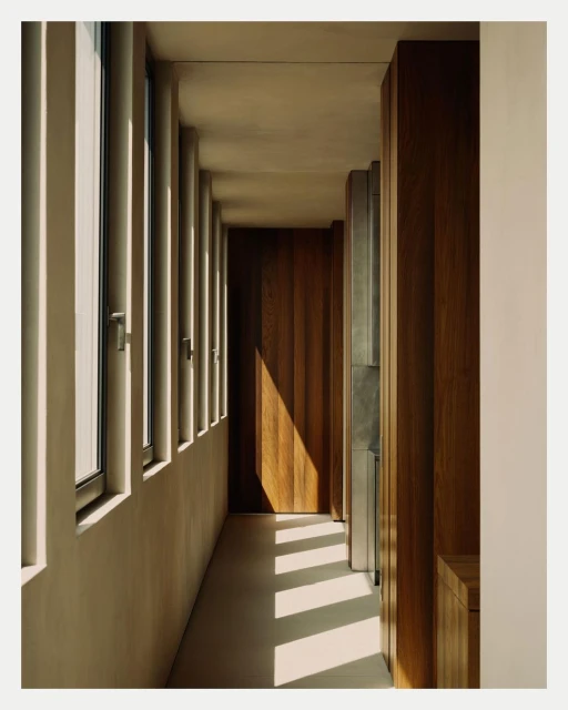 Interior shot of a hallway with natural light streaming through tall windows and reflecting off a wood door, creating a warm, architectural scene.