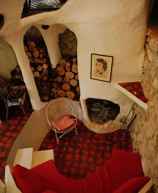 Unique interior design featuring a red sofa, wicker chair, and firewood, accented by an art piece, all within a cave-like room structure.