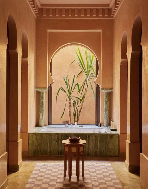 Luxurious peach-toned bathroom features a tub, marble accents, arched doorway, potted plant, geometric floor tiles, and pedestal table.