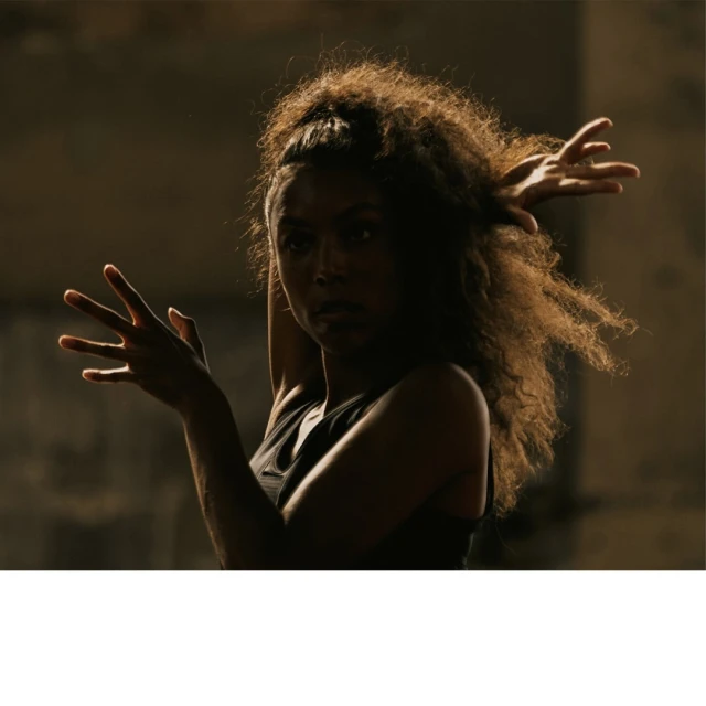 Woman in a tank top with curly hair poses in front of a textured grey wall. The lighting is dramatic and directional.
