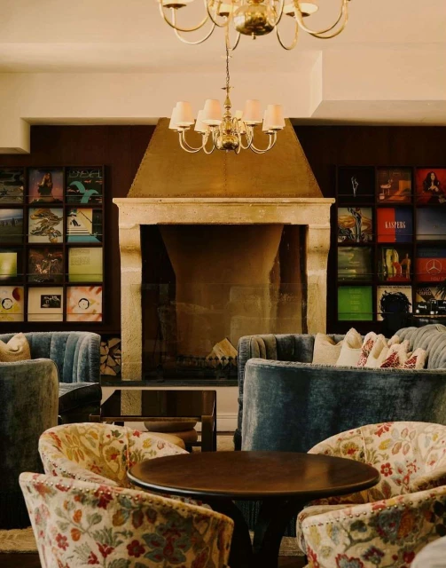 Interior of a hotel lounge area with floral armchairs, blue velvet sofas, a fireplace, and a decorative chandelier. Warm, inviting space.