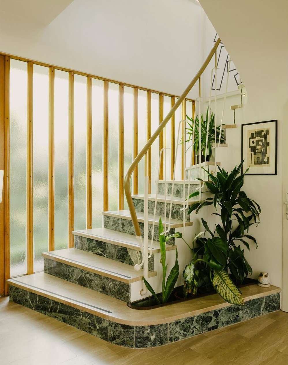 Indoor shot features a staircase with green marble trim, lush potted plants, and natural light filtering through vertical wood slats.