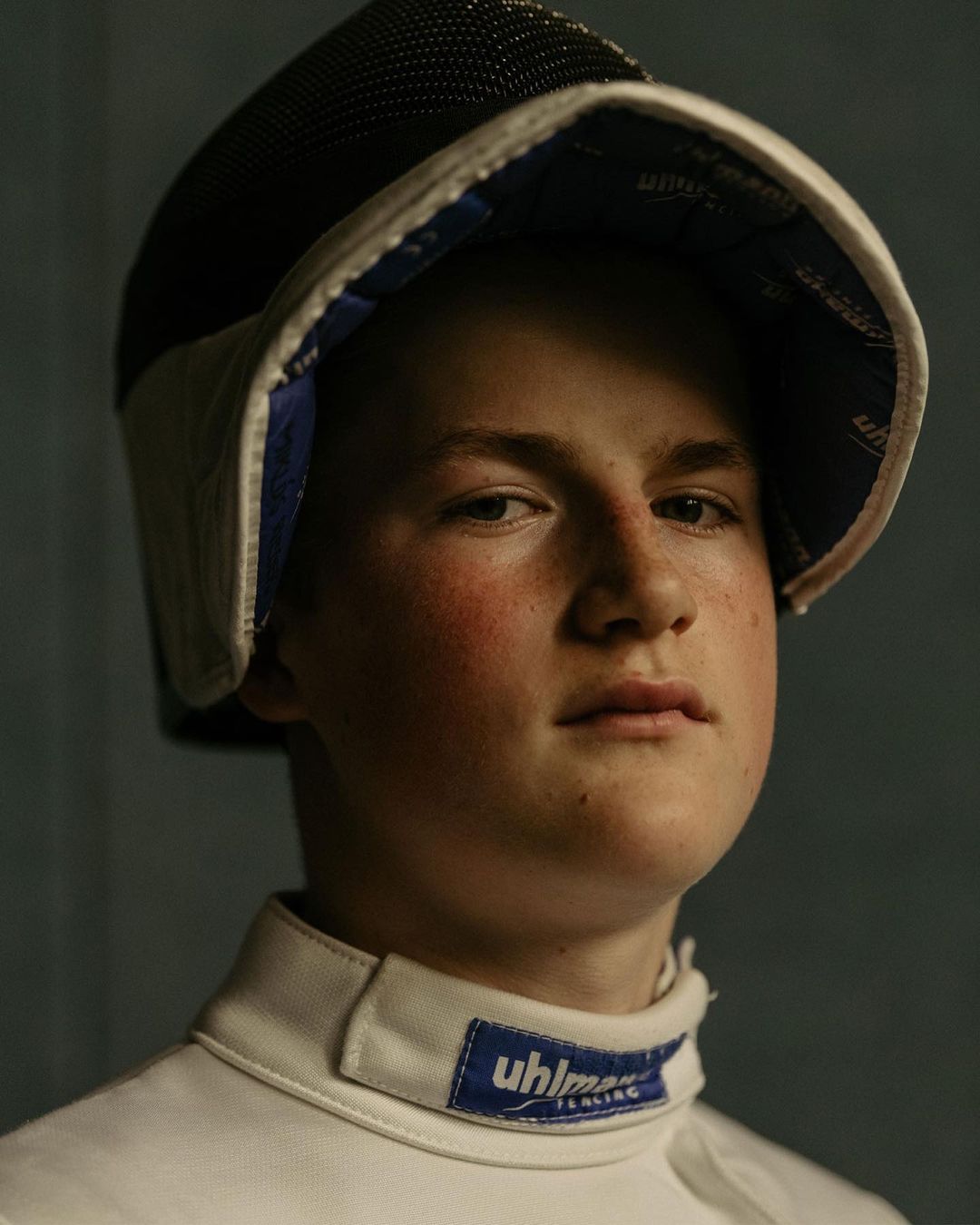 Close-up studio portrait of a young adult male wearing a fencing mask and protective gear, looking at the camera against a grey background.