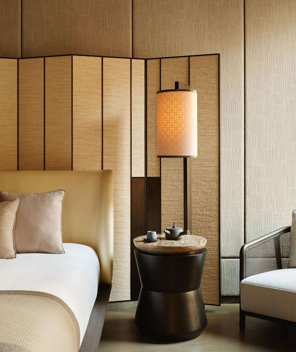 Interior of a hotel room featuring a bed with a beige headboard, an adjacent end table with a lamp, and a chair, all in neutral tones.