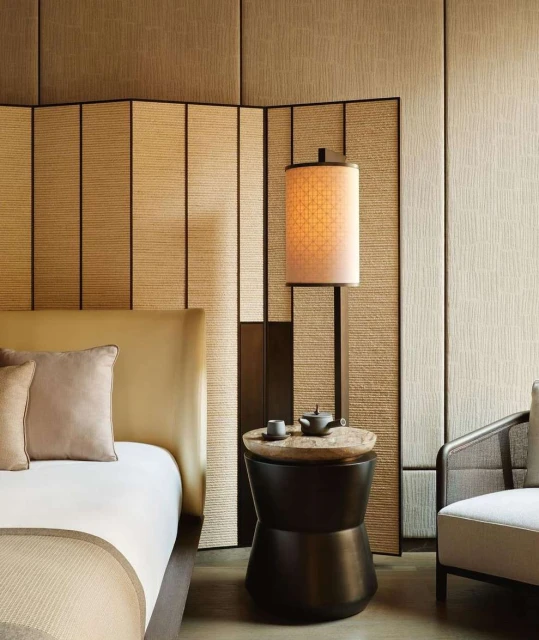 Interior of a hotel room featuring a bed with a beige headboard, an adjacent end table with a lamp, and a chair, all in neutral tones.