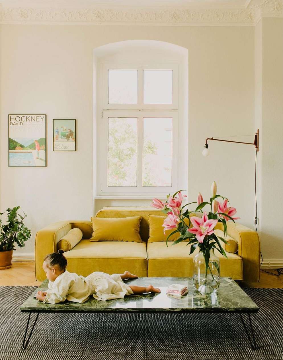 Child lying on a green marble coffee table in front of a yellow sofa, with wall art and a houseplant in a bright living room.