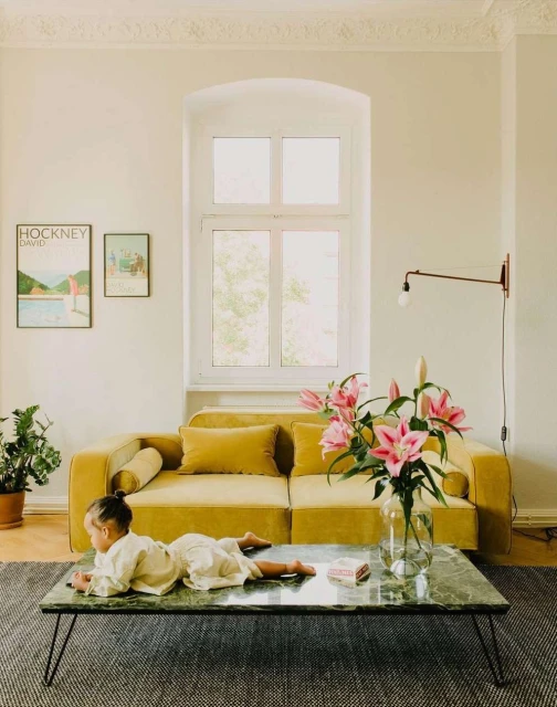 Child lying on a green marble coffee table in front of a yellow sofa, with wall art and a houseplant in a bright living room.