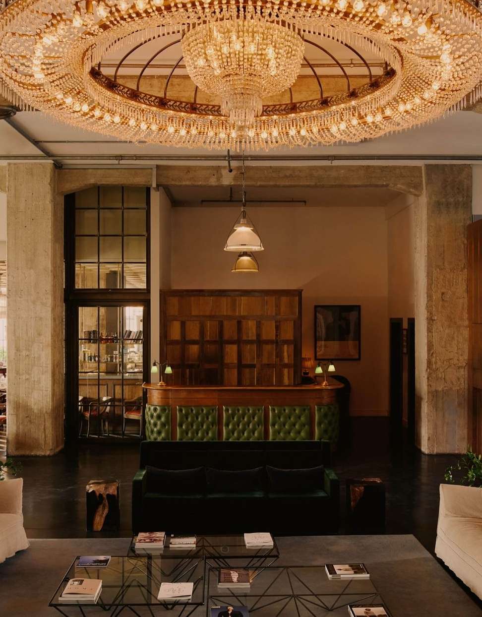 Opulent interior with a large crystal chandelier hanging above a green velvet sofa, glass tables, and minimal decor in a spacious room.