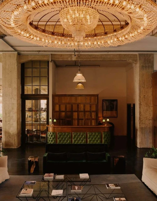 Opulent interior with a large crystal chandelier hanging above a green velvet sofa, glass tables, and minimal decor in a spacious room.