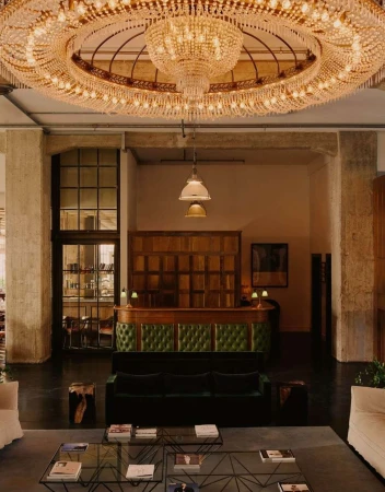 Opulent interior with a large crystal chandelier hanging above a green velvet sofa, glass tables, and minimal decor in a spacious room.