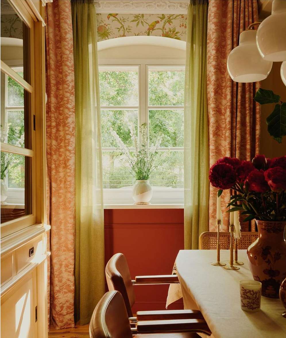 Dining room interior with a white table, chairs, a floral arrangement, curtains, and natural light from a large window.