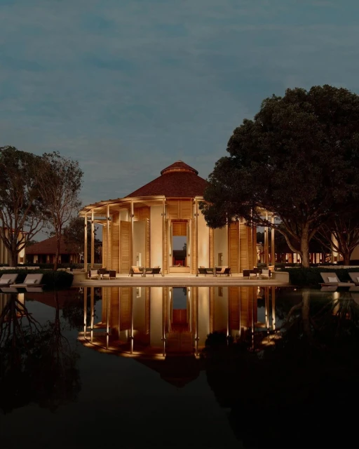 Exterior of a modern building with a brown roof reflecting in a dark pool of water surrounded by trees at twilight.