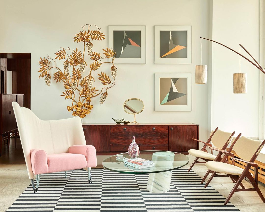 Living room interior with pink wingback chair, wooden sideboard, round glass coffee table, striped rug, and decorative gold wall art.