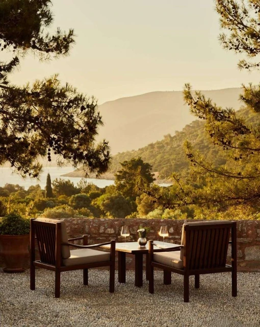 Outdoor dining scene with two wooden chairs and a small table set with drinks, overlooking a lush landscape and distant hills at dusk.