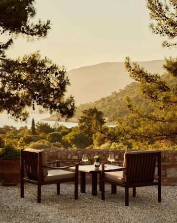 Outdoor dining scene with two wooden chairs and a small table set with drinks, overlooking a lush landscape and distant hills at dusk.