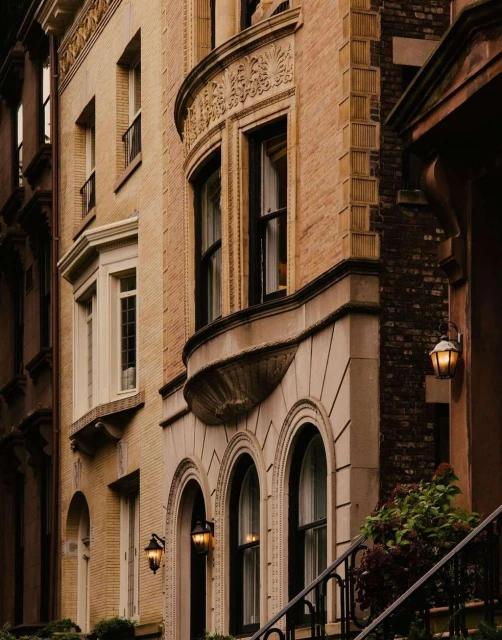 Exterior of residential brownstone buildings with detailed architecture and illuminated wall sconces lining facades in an urban environment.