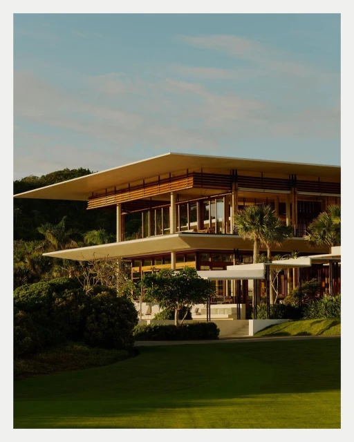 Exterior view of a multi-level modern home with flat roofs and glass walls, surrounded by green lawns and trees under a clear blue sky.