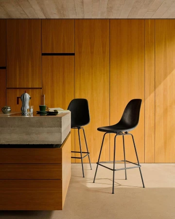 Minimalist kitchen interior featuring two black counter stools, a concrete countertop, wooden cabinets, and natural lighting.