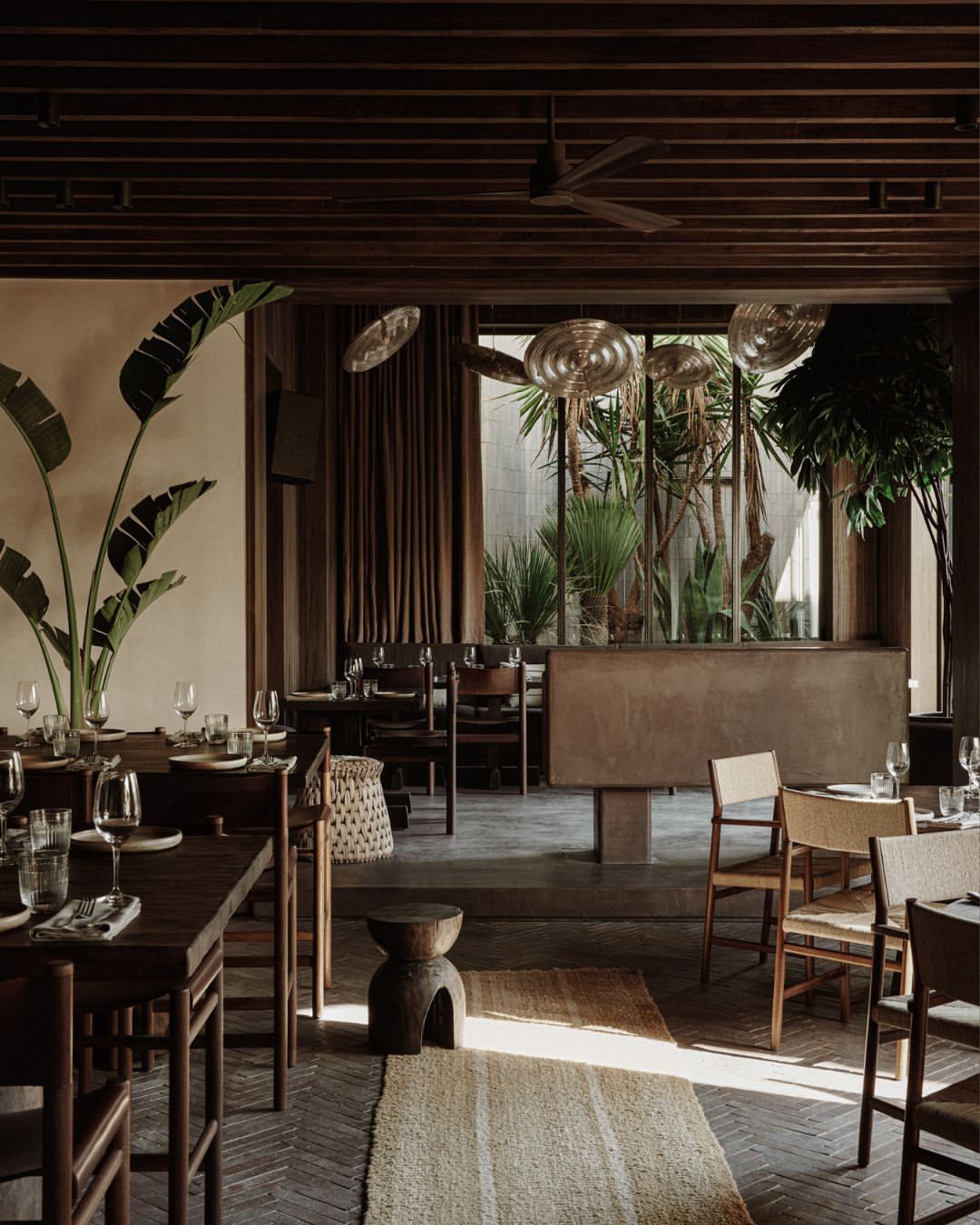 Interior of a restaurant featuring tables set with glassware and tableware, potted plants, and wooden furniture in warm lighting.