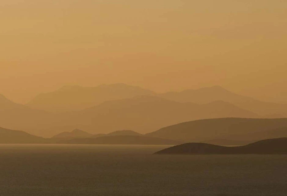 Scenic view of a calm body of water reflecting the warm, golden light of the sky, with layered, silhouetted hills in the background.