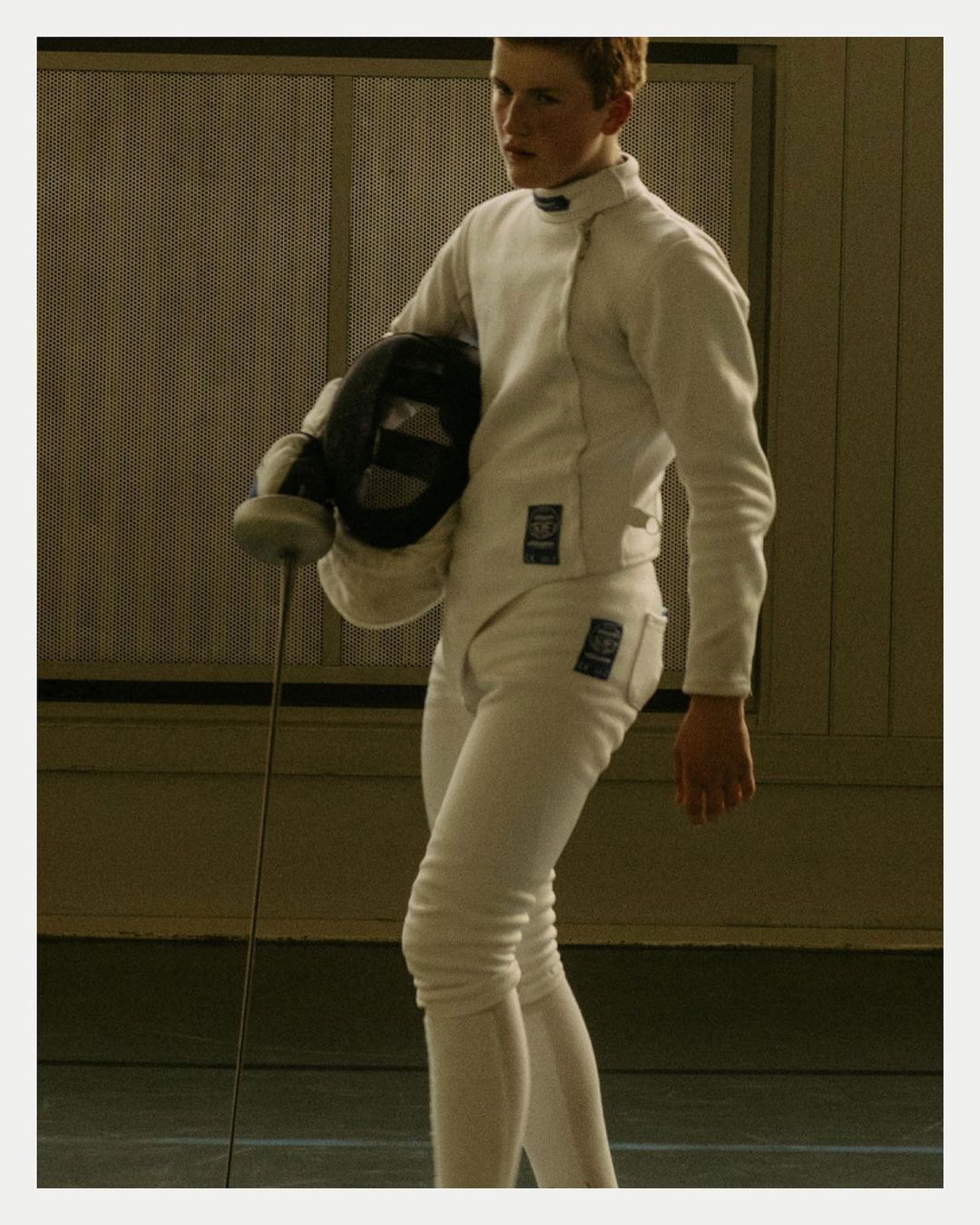 A fair-skinned fencer in white uniform holds a mask and foil indoors with a neutral facial expression in a medium shot, athletic and focused.