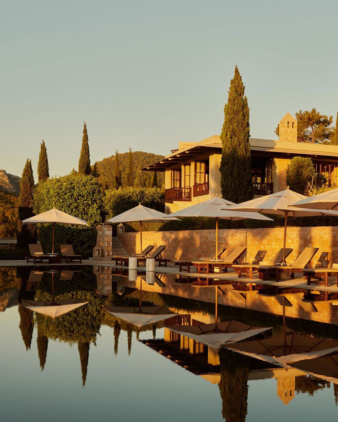 Exterior of a luxury resort with sun loungers and umbrellas reflected in a calm pool at sunset, creating a serene and inviting scene.