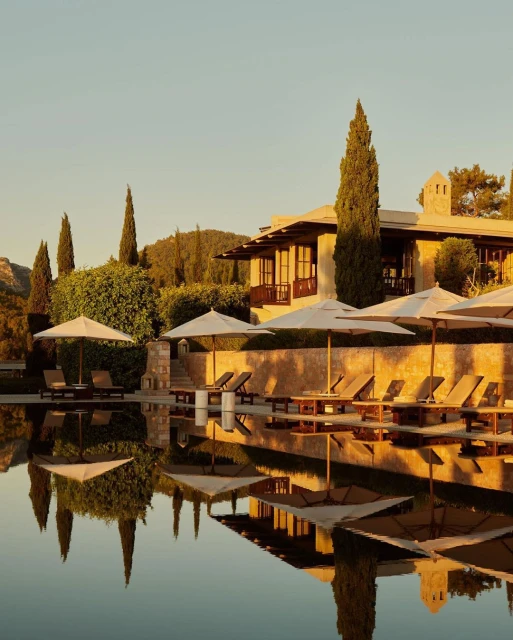 Exterior of a luxury resort with sun loungers and umbrellas reflected in a calm pool at sunset, creating a serene and inviting scene.