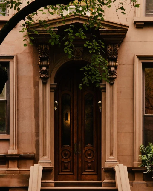 Brownstone facade with arched wooden double doors and decorative stonework, framed by an overhanging tree branch with green leaves.