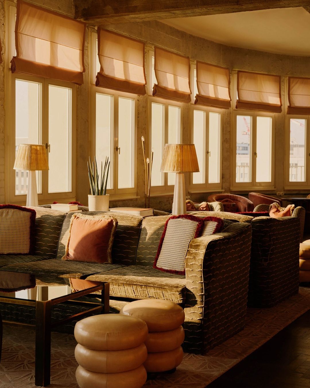 Cozy interior with patterned sofas, soft pillows, and a table with stacked ottomans, all lit by warm light from large windows.