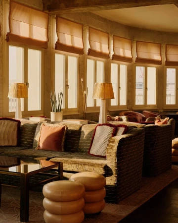 Cozy interior with patterned sofas, soft pillows, and a table with stacked ottomans, all lit by warm light from large windows.