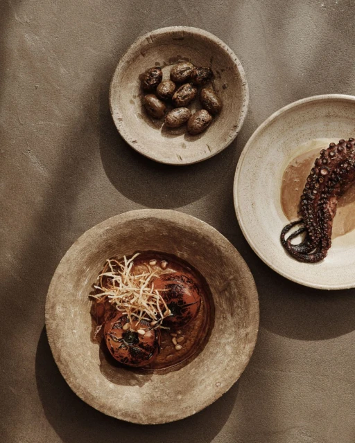 Overhead shot of three plates with grilled octopus, tomatoes and olives. The plates are resting on a neutral brown background.