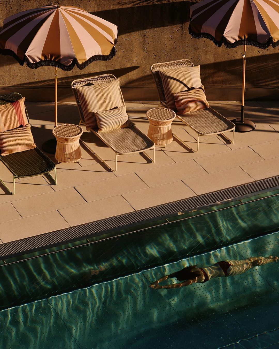 A woman swims in a pool with lounge chairs, side tables, and striped parasols beside it, capturing a tranquil, luxurious outdoor scene.