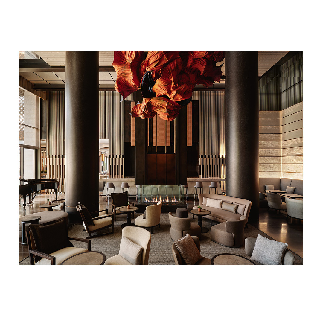 Hotel lobby lounge area with comfortable seating arrangement, modern fireplace, piano, and a dramatic floral chandelier.
