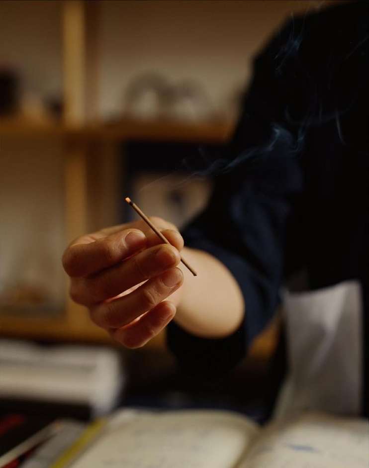 Close-up of a hand holding a burning incense stick with smoke rising indoors, set against a blurred background with wooden shelves.