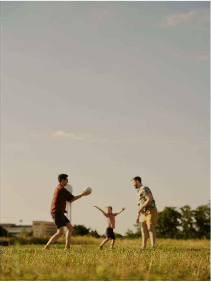 Three people playing with a ball in a grassy field with a building in the distance under a bright sky, family outdoor activity.