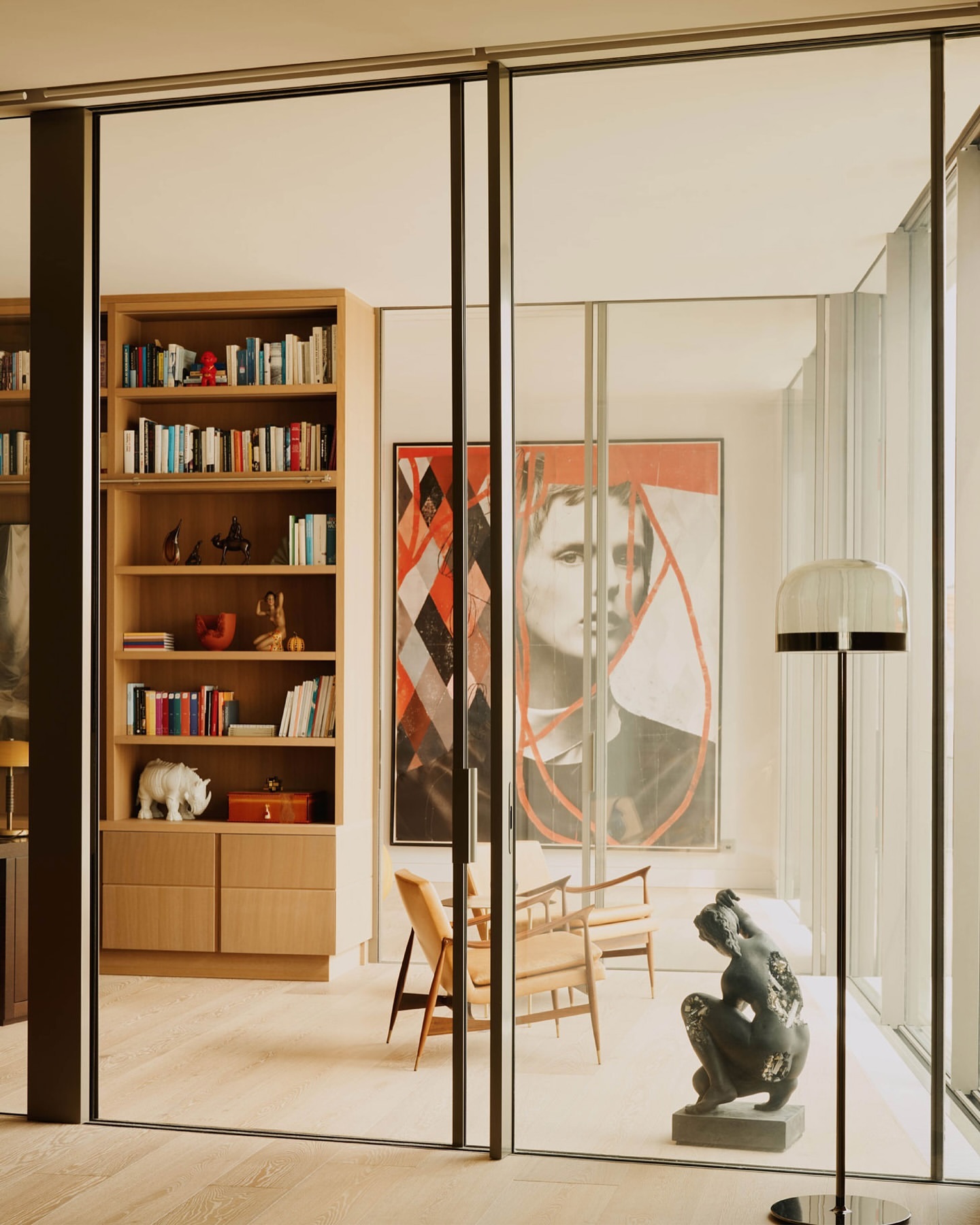 Home interior with a glass wall separating two rooms, a bookcase, artwork, sculptures, armchairs, and a floor lamp.