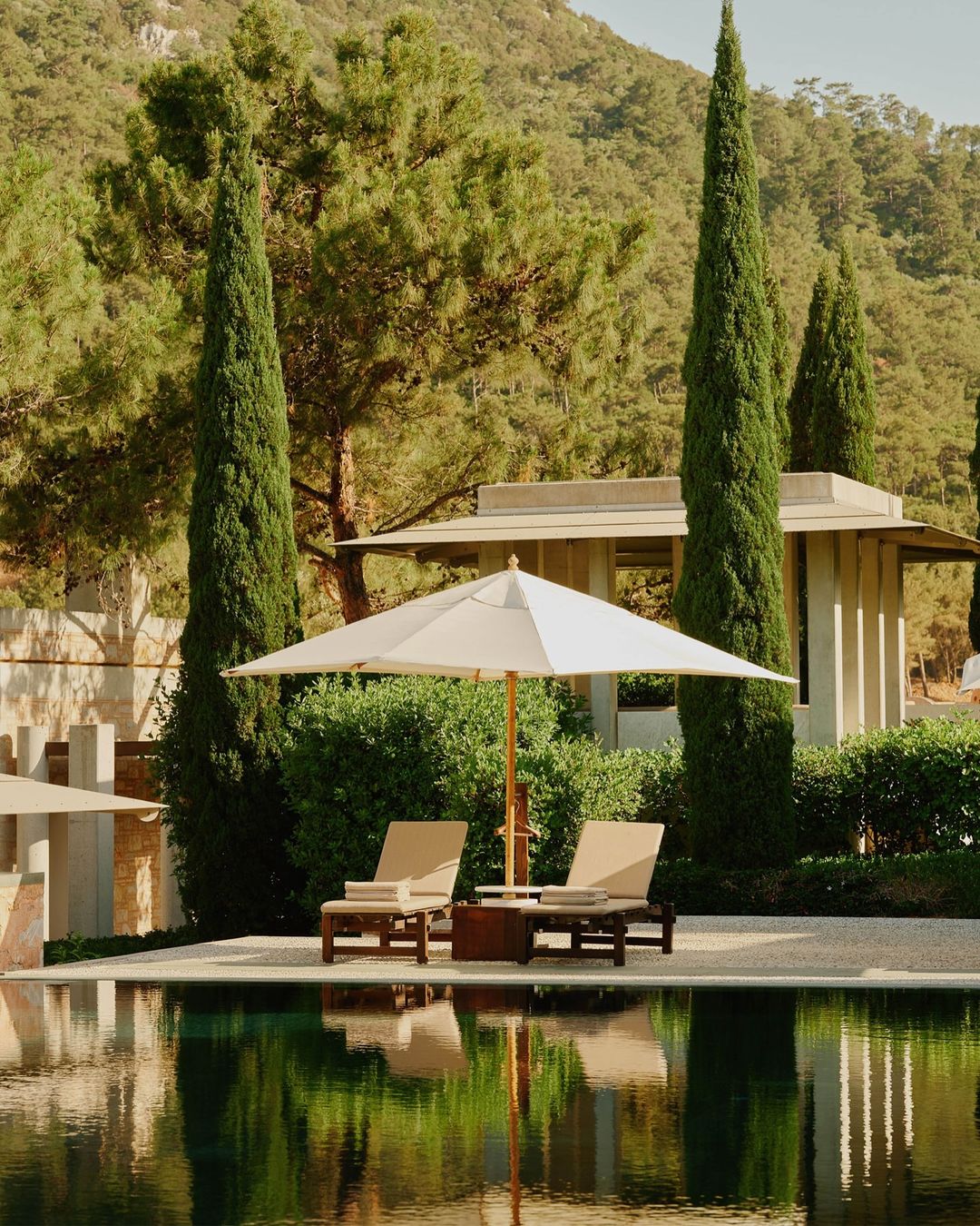 Outdoor relaxation area featuring lounge chairs under a sun umbrella by a reflective pool, surrounded by green trees and manicured gardens.