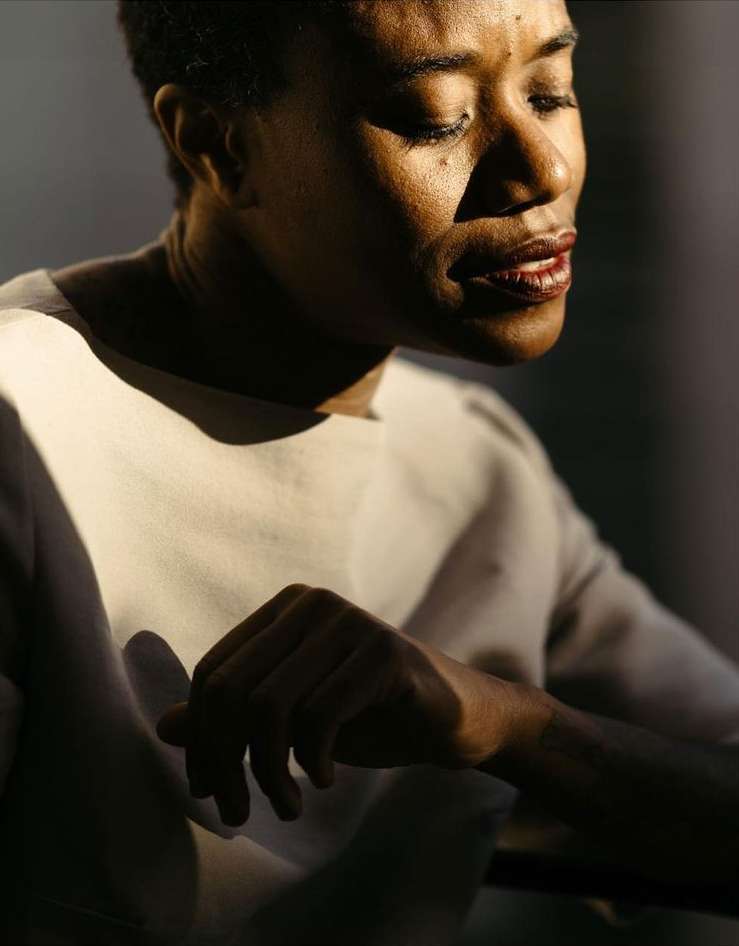A dark-skinned adult female gazes downwards in a close-up shot. She wears a cream top with shadows playing across her face and hand in a soft lighting.