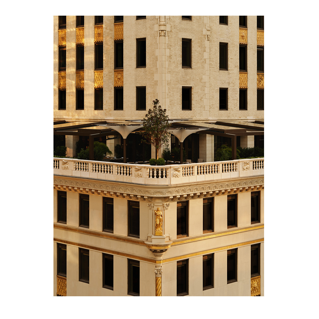 View of a tan-colored, vintage building featuring a rooftop terrace with a small tree, decorative gold accents, and symmetrically aligned windows.
