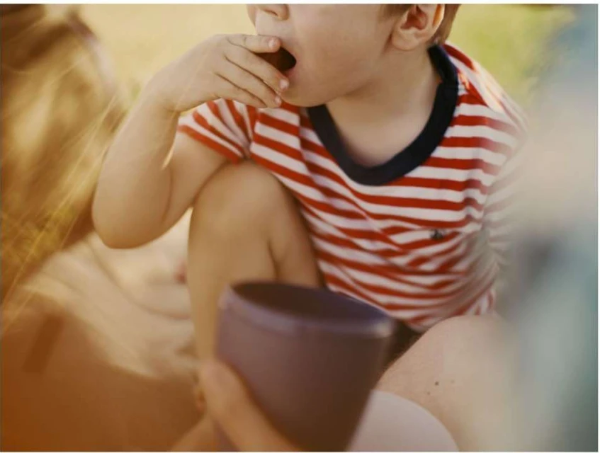 Young child with red and white striped shirt eats while seated in sunny outdoor setting with a purple bowl on their lap.